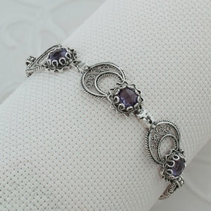 May include: A silver bracelet with three purple gemstones set in filigree designs. The bracelet is made of delicate, intricate wirework.