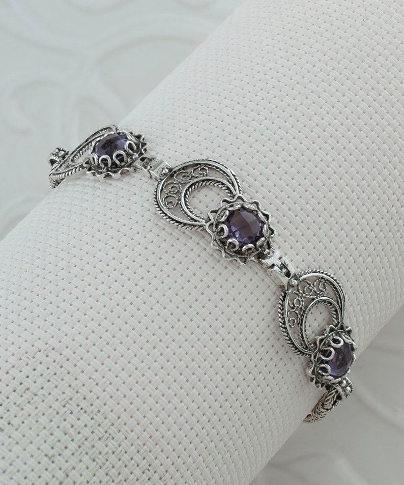 May include: A silver bracelet with three purple gemstones set in filigree designs. The bracelet is made of delicate, intricate wirework.