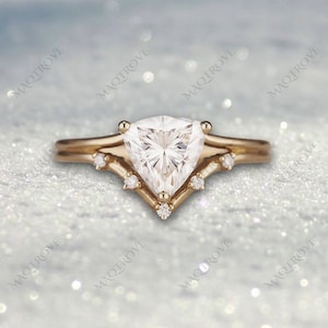 Könnte beinhalten: Ein goldener Verlobungsring mit einem dreieckigen Diamanten als Mittelstück und kleineren Diamanten auf der Ringschiene. Der Ring liegt auf einem weißen, glitzernden Hintergrund.