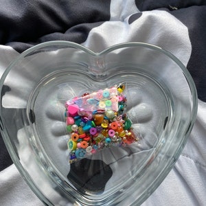 May include: A clear heart-shaped glass bowl containing a plastic bag filled with colorful beads. The beads are a variety of shapes and sizes, including round, square, and star-shaped. The beads are in a rainbow of colors, including red, orange, yellow, green, blue, purple, and pink.