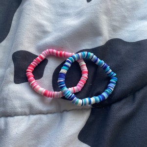 May include: Two colorful bracelets made of stacked, round, plastic beads. One bracelet is pink, white, and blue. The other bracelet is blue, white, and light blue.
