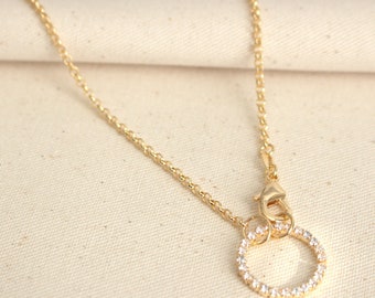 Può includere: Una collana con catena d'oro con un piccolo ciondolo rotondo. Il ciondolo è in oro e ha un cerchio di pietre trasparenti attorno al bordo.