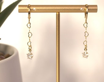 May include: Gold hoop earrings with a delicate chain and a star-shaped crystal charm. The earrings are displayed on a gold stand.