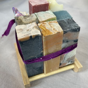 May include: A set of nine different colored bars of soap arranged in a cube shape on a wooden stand. The soap bars are tied together with a purple ribbon.