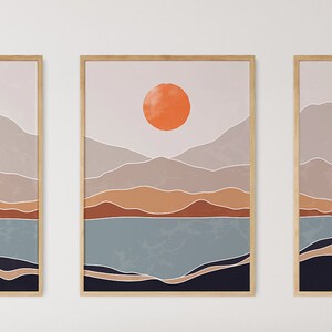 May include: A set of three abstract landscape prints featuring a mountain range, a lake, and a large orange sun. The prints are framed in natural wood.