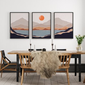 May include: Three framed abstract landscape prints with a minimalist style. The prints feature a mountain range with a lake in the foreground and a large orange sun in the top center of the middle print.