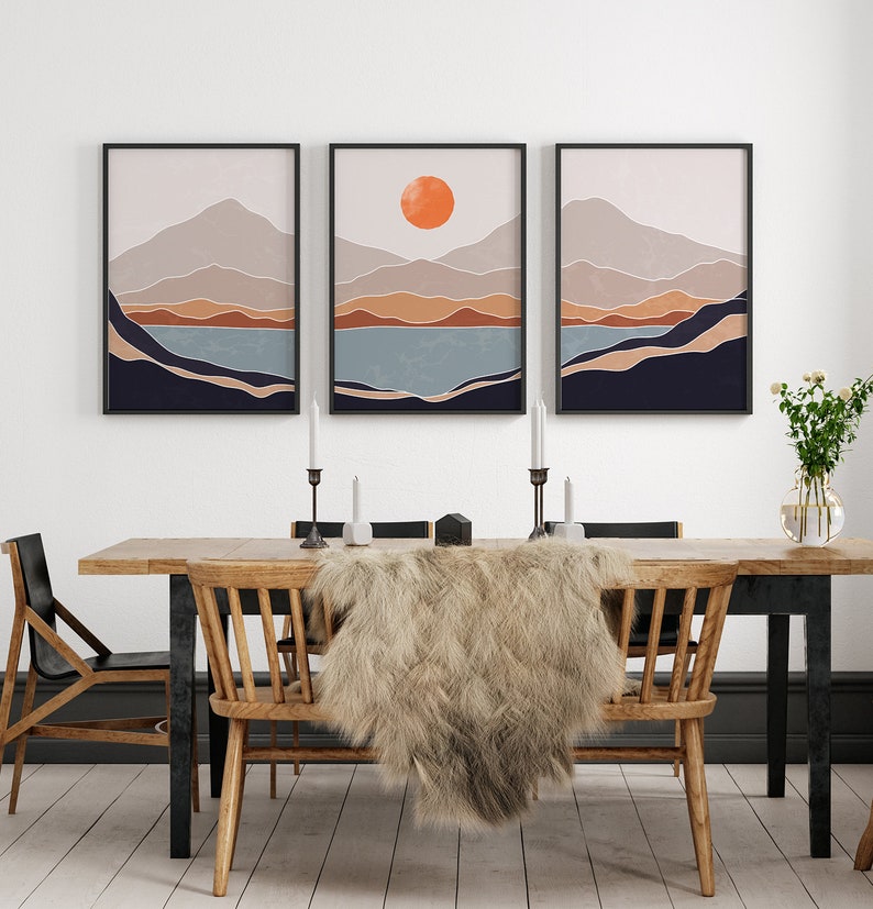 May include: Three framed abstract landscape prints with a minimalist style. The prints feature a mountain range with a lake in the foreground and a large orange sun in the top center of the middle print.
