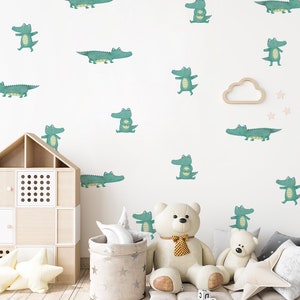 Può includere: Una parete bianca decorata con adesivi murali di alligatori verde acqua in varie pose. Una mensola a forma di casa in legno, un cesto a motivi di stelle e animali di peluche, tra cui un orsacchiotto, sono in primo piano, creando un tema giocoso per la cameretta.