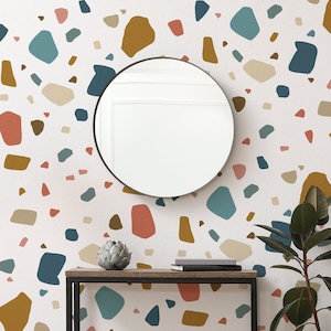 Può includere: Uno specchio rotondo con una cornice bianca è appeso a una parete con una carta da parati a motivo terrazzo. La carta da parati presenta una varietà di forme e colori, tra cui marrone, beige, blu e rosa. Un tavolo da console marrone scuro con un telaio in metallo nero si trova di fronte al muro. Una piccola pianta in vaso si trova sul tavolo, insieme a due libri.