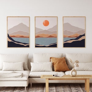 May include: Three abstract landscape prints with a minimalist style. Each print features a mountain range with a lake in the foreground and a sunset in the background. The prints are framed in a light wood frame.