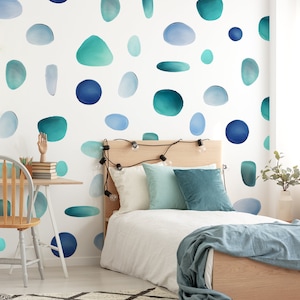 Op de afbeelding: Slaapkamer met een wit bed, houten hoofdbord en een bureau. De witte muur is versierd met blauwe en turquoise aquarel vormen. Een blauwe deken en kussens geven het bed kleur. Een houten stoel en bureau staan in de hoek.