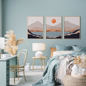 May include: Three abstract landscape prints in a minimalist style with a muted color palette of blue, brown, and orange. The prints feature a mountain range, a body of water, and a sun. The prints are framed in a simple wooden frame.
