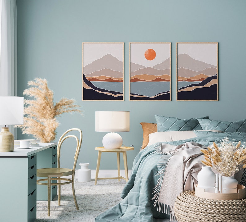 May include: Three abstract landscape prints in a minimalist style with a muted color palette of blue, brown, and orange. The prints feature a mountain range, a body of water, and a sun. The prints are framed in a simple wooden frame.