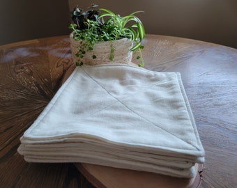 Può includere: Un mucchio di strofinacci di cotone bianco con una texture sottile. Gli strofinacci sono piegati a metà e hanno un bordo cucito. Gli strofinacci sono su una superficie di legno.