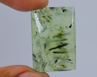 May include: A green and black gemstone cabochon with a rectangular shape. The stone has a translucent appearance with black inclusions.