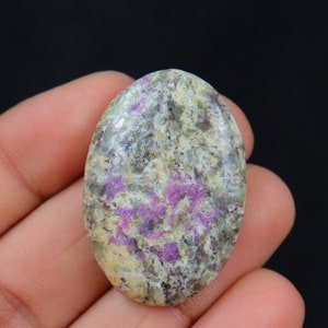 Peut inclure: Une pierre précieuse ovale avec un motif moucheté de teintes vertes, grises et violettes. La pierre polie est tenue dans une main sur un fond noir, mettant en valeur ses couleurs et textures naturelles. Cette pierre convient à la création de bijoux.