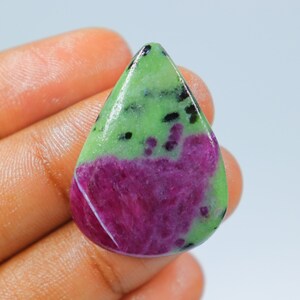 Puede incluir: Un cabujón de piedra preciosa verde y rosa en forma de lágrima con inclusiones negras. La piedra preciosa se sostiene en una mano.