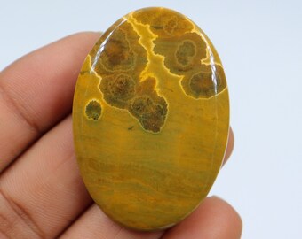 May include: A yellow and brown oval cabochon gemstone with a unique pattern of dark brown and green spots. The gemstone is smooth and polished.