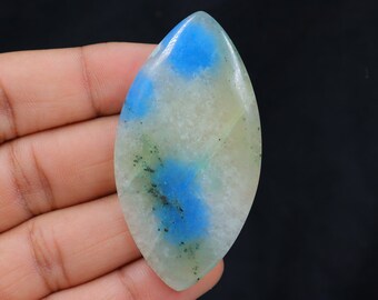 May include: A polished, elongated gemstone with a leaf-like shape. The stone features a blend of colors, including light blue, white, and green, with darker speckles. The gemstone is held in a hand against a black background.