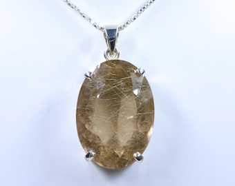 May include: A silver necklace with an oval, golden-brown gemstone pendant. The pendant is held in place by four silver prongs and features internal golden-colored inclusions. The chain is a delicate, textured silver. The background is a plain white.