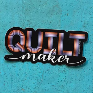 May include: A black sticker with the words "QUILT maker" in white script. The word "QUILT" is in large, retro-style letters with orange, purple, and pink stripes. The sticker has a turquoise background.