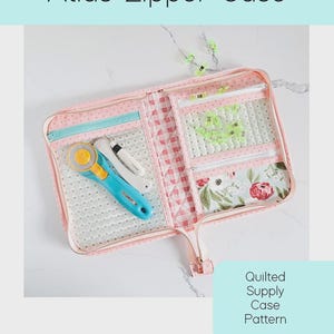 May include: A pink quilted Atlas Zipper Case with a light blue zipper and accents. The case contains a rotary cutter and other sewing tools. The case is labeled as a quilted supply case pattern.