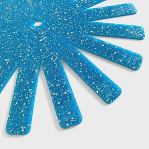 May include: A close-up of a blue, star-shaped object with glitter. The object has a central hole and radiating, rectangular arms. The arms are speckled with silver and iridescent glitter.