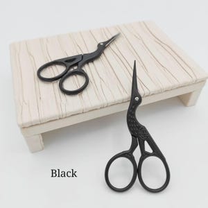 May include: Two black crane-shaped embroidery scissors. The scissors are made of metal and have a bird-like design. The text "Black" is visible in the image. The scissors are on a light-colored, textured surface.