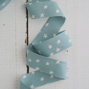 May include: A roll of light blue fabric ribbon with a repeating pattern of white stars. The ribbon is curled and draped, showcasing the star design. The background is a white wooden surface.