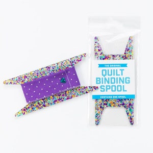 May include: A purple and white polka dot fabric is wrapped around a clear plastic spool with colorful confetti-like speckles. The spool is labeled "The Original Quilt Binding Spool" and "Contains One Spool".