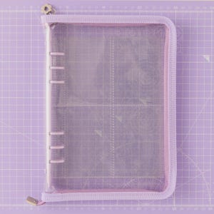 May include: A clear, zippered binder with a lavender fabric trim and a silver zipper pull. The binder has a ring mechanism and is set against a lavender grid background, suggesting its use for organizing papers or crafts.