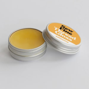 May include: Open tin of Sew Fine Thread Gloss with a yellow balm inside, next to the lid. The lid is orange with white text that reads "Sew Fine Thread Gloss" and "satsuma". The tin is silver.