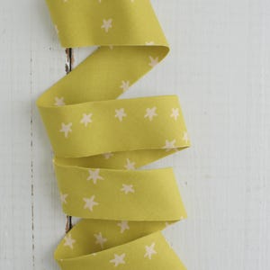 May include: A length of yellow-green fabric ribbon with a repeating pattern of small, light pink stars. The ribbon is curled and folded, creating a zig-zag pattern. The background is a white, weathered wooden surface.