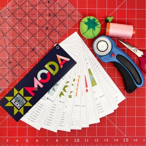 May include: A close-up of a Moda Modern Building Blocks pattern book with a blue, red, and green cover. The book is open to a page with a grid of squares and a variety of colorful fabric squares. The book is on a red cutting mat with a rotary cutter, a pink thread spool, and a green pincushion.