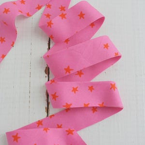 May include: A length of pink fabric ribbon with a repeating pattern of orange stars. The ribbon is folded and arranged in a flowing, curved shape against a white wooden surface. The fabric has a matte finish.