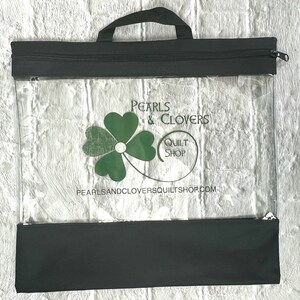 May include: Black zippered tote bag with a clear plastic front panel. The bag has a black handle and a green four-leaf clover logo with the text "Pearls & Clovers Quilt Shop" and "pearlsandcloversquiltshop.com".