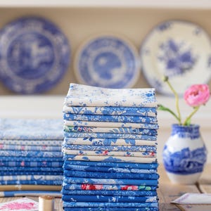 May include: A tall stack of folded fabric squares featuring various shades of blue and white floral, botanical, and geometric patterns. The stack is arranged on a wooden surface alongside a small wooden spool, scattered buttons, and gold scissors.