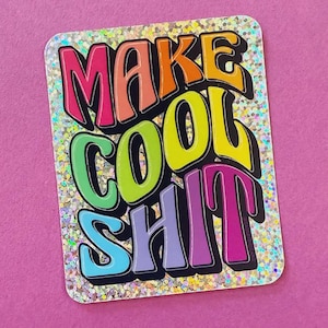 May include: A rectangular sticker with the words "MAKE COOL SHIT" in colorful bubble letters. The letters are outlined in black and have a glittery background. The sticker is on a pink background.