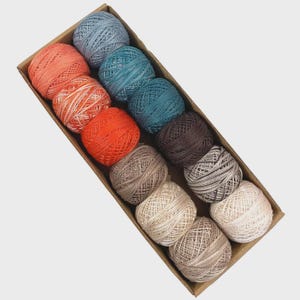 May include: A rectangular cardboard box filled with twelve balls of colorful yarn. The yarn balls are in shades of orange, blue, teal, brown, and beige. The yarn is tightly wound and appears to be cotton or a similar fiber. The box is angled, showing the yarn in a neat arrangement.