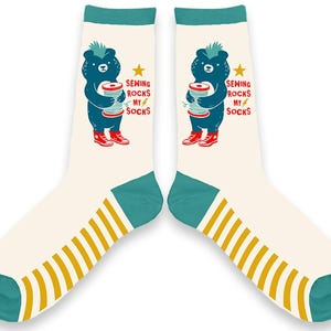 May include: A pair of white socks with teal stripes and a blue bear graphic. The bear is holding a spool of thread and the text "Sewing Rocks My Socks" is printed on the socks.