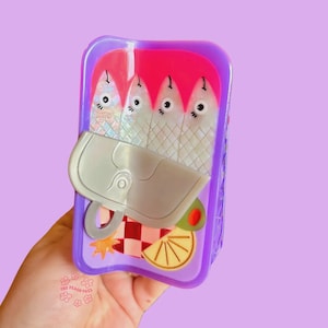 May include: A novelty sardine can-shaped accessory in a rectangular purple case. The design features fish, a can opener, an olive, and a lemon slice. The can is held in a hand against a purple background.