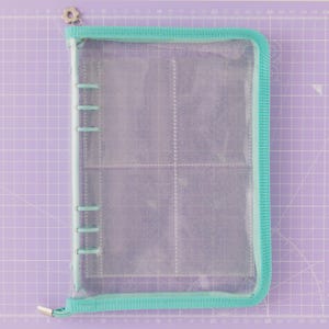 May include: A clear, zippered binder with a teal trim and a silver zipper pull. The binder has multiple clear pockets inside, designed for storing small items. The background is a purple cutting mat with grid lines.