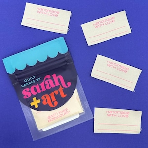 May include: A package of quilt labels by Sarah + Art, with several individual labels scattered around. The labels are rectangular, with the words "Handmade WITH LOVE" printed in pink. The package has a navy blue background with a teal scalloped top.