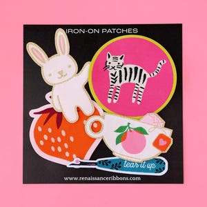 May include: A set of five iron-on patches featuring a white bunny, a striped cat, a strawberry, a teacup with a peach, and a blue tool with the text "tear it up". The patches are on a black background with the text "IRON-ON PATCHES" and "www.renaissanceribbons.com".