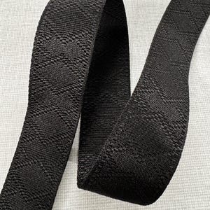 May include: Black woven strap with a repeating geometric pattern. The strap is made of a durable material and is suitable for various crafting and sewing projects. The strap is shown against a light gray background.