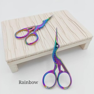 May include: Two rainbow-colored embroidery scissors with a bird design. The scissors are displayed on a small, light-colored wooden platform. The word "Rainbow" is printed in black at the bottom of the image.