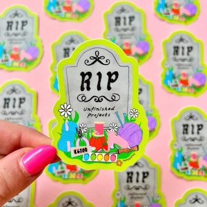 May include: A sticker with a tombstone design that reads "RIP Unfinished Projects." The sticker features illustrations of yarn, knitting needles, and other craft supplies, with a bright green border and a pink background.