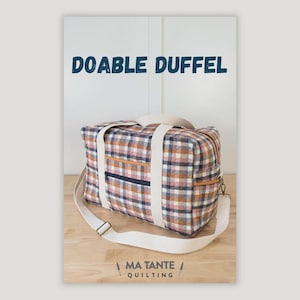 May include: A plaid duffel bag with a cream-colored shoulder strap and handles. The bag features a zipper pocket and orange trim. The text "DOABLE DUFFEL" is printed in blue above the bag. The brand name "MA TANTE QUILTING" is printed at the bottom.