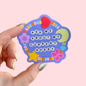 May include: A purple sticker with a beaded border and colorful charms. The sticker says "Ask Me About My Current Hobby" in white letters.