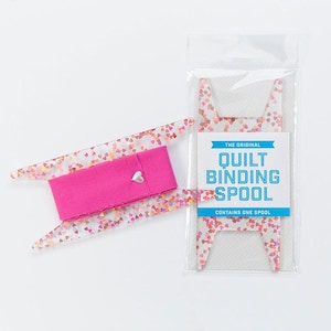 May include: A clear plastic spool with a pink fabric ribbon and a silver heart charm. The spool is decorated with a pink and orange glitter heart pattern. The packaging says "The Original Quilt Binding Spool. Contains one spool."
